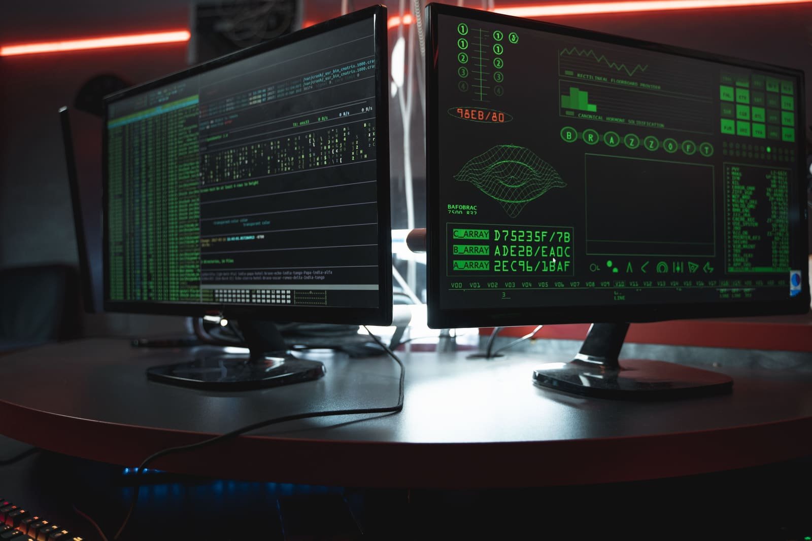 Deux écrans de supervision affichant du code et des courbes vertes dans une salle éclairée de rouge